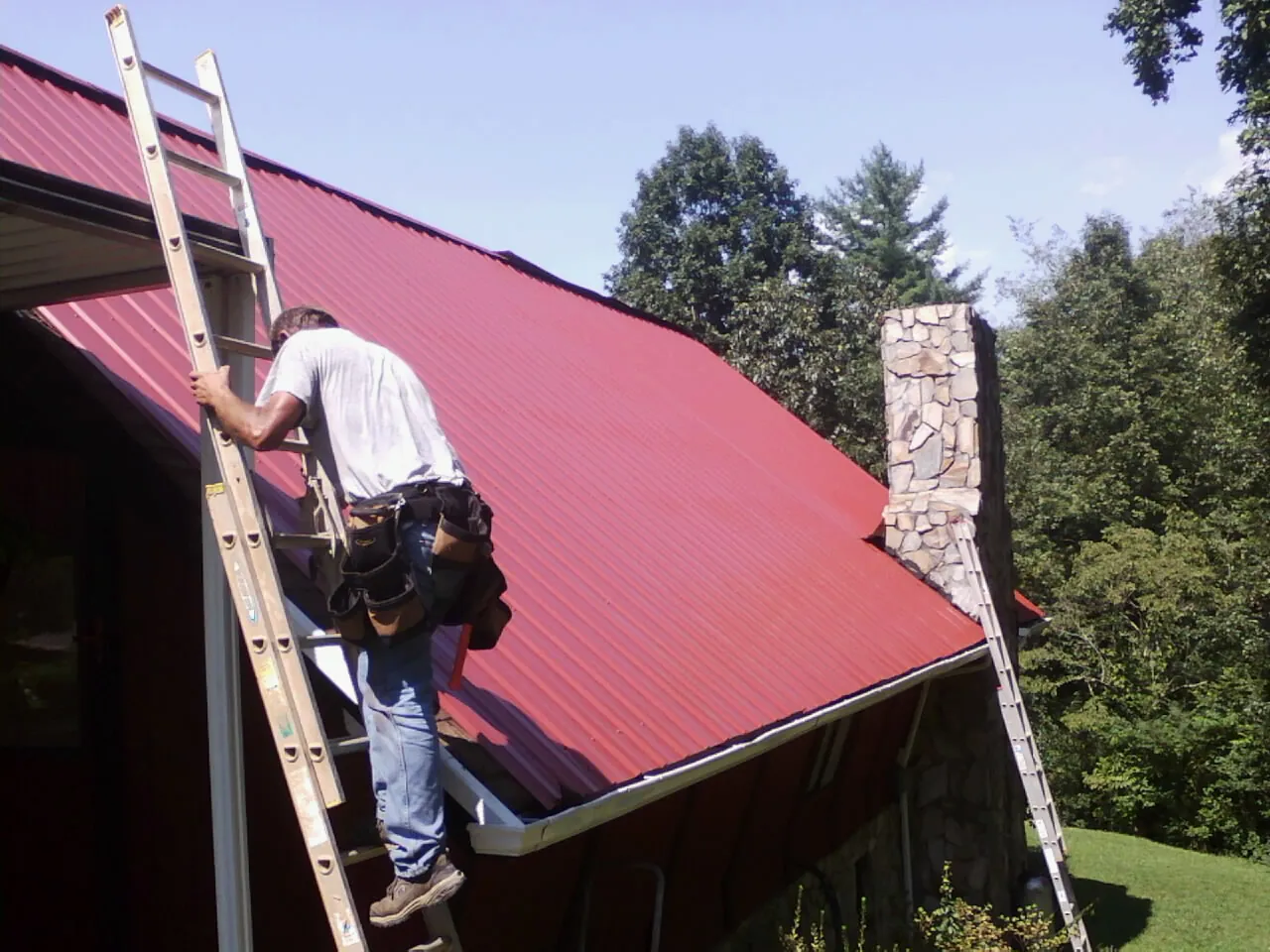 Roofer on a ladder beside a red metal roof and stone chimney — Soffit & Fascia Repair by Roofing Brothers in Bloomfield Hills